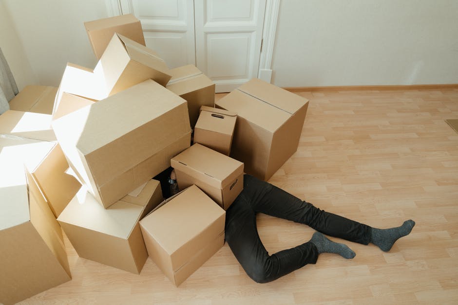 A person wearing black pants and grey socks is partially hidden underneath a stack of packed cardboard moving boxes inside a room with light wooden flooring and cream-colored walls. The boxes vary in size, some are plain cardboard with no labels, and are arranged haphazardly, with one large box leaning against a closed white door with decorative paneling. The boxes are likely filled with household items and are prepared for packing and moving. There are some plastic bottles and packing materials visible among the boxes. The scene captures the loading process during home relocation, reflecting the work of a professional removals service such as Man and Van Harrow as they prepare furniture transport and assist with packing and moving logistics.