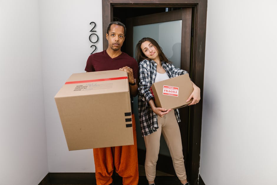A person wearing black pants and grey socks is partially hidden underneath a stack of packed cardboard moving boxes inside a room with light wooden flooring and cream-colored walls. The boxes vary in size, some are plain cardboard with no labels, and are arranged haphazardly, with one large box leaning against a closed white door with decorative paneling. The boxes are likely filled with household items and are prepared for packing and moving. There are some plastic bottles and packing materials visible among the boxes. The scene captures the loading process during home relocation, reflecting the work of a professional removals service such as Man and Van Harrow as they prepare furniture transport and assist with packing and moving logistics.