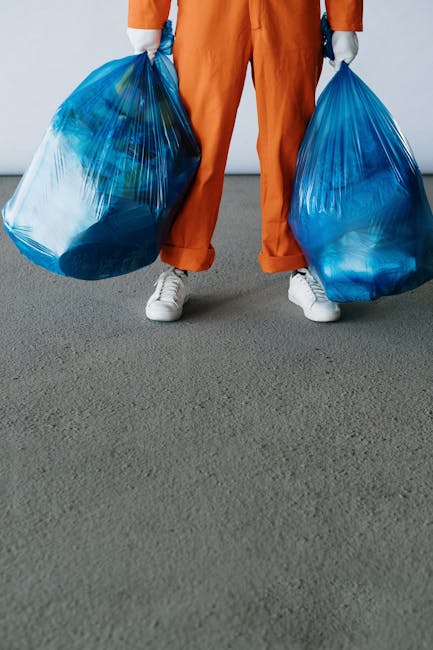 A person wearing orange overalls and white sneakers is standing inside a property, holding two large blue plastic garbage bags filled with waste materials, likely related to house removals or clearing. The bags contain various discarded items such as cardboard boxes, packaging, and possibly textiles, and are being carried to an exterior area or a waste collection point. The scene occurs on a grey carpeted floor near a doorway, with the background showing a plain, light-colored interior wall. The image captures the process of packing or disposing of bulky waste as part of a home relocation or furniture transport task, reflecting the types of cleanup activities managed by professional removals services like Man and Van Harrow, in accordance with local council disposal rules for Harrow.