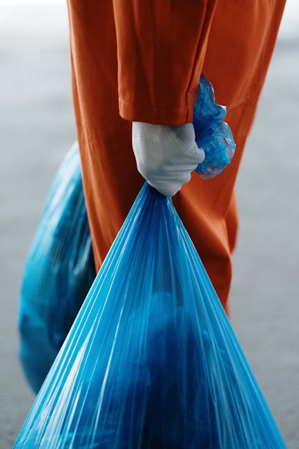 Inside a residential property, a person wearing orange work overalls and white gloves is seen holding a blue plastic bag filled with bulky waste material, likely relating to house removals or clearance. The person is standing near an open doorway with a view of the interior floor and furniture, including cardboard boxes, a wooden chair, and packaging materials such as plastic wrap and fabric coverings, indicating a packing or packing-up process. The setting is illuminated by natural light, highlighting the space temporarily used for sorting and loading items. Nearby, a van with a partially visible rear area is positioned outside on a paved driveway or street, where additional furniture pieces or boxes might be loaded for transport. The scene represents an organized loading process typical of home relocation or rubbish removal services, with Man and Van Harrow providing the logistics of furniture transport and disposal according to local house removals standards and regulations.