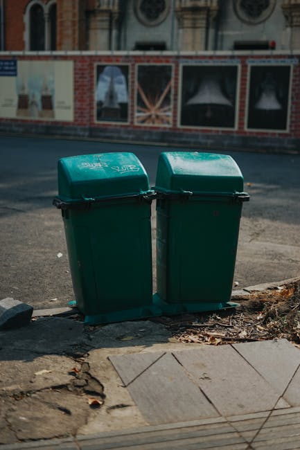 A person wearing orange overalls and white sneakers is standing inside a property, holding two large blue plastic garbage bags filled with waste materials, likely related to house removals or clearing. The bags contain various discarded items such as cardboard boxes, packaging, and possibly textiles, and are being carried to an exterior area or a waste collection point. The scene occurs on a grey carpeted floor near a doorway, with the background showing a plain, light-colored interior wall. The image captures the process of packing or disposing of bulky waste as part of a home relocation or furniture transport task, reflecting the types of cleanup activities managed by professional removals services like Man and Van Harrow, in accordance with local council disposal rules for Harrow.