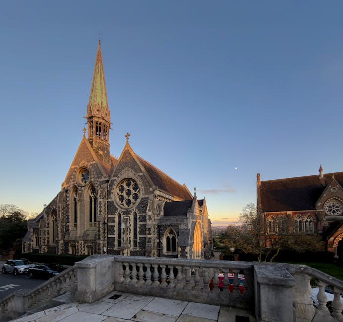 This image shows a street scene outside a historic church with a tall, pointed spire, situated in a residential area during daylight hours with clear blue skies. The church is built from stone and fea