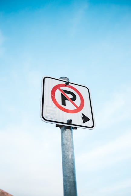 A close-up view of a white rectangular sign mounted on a metal pole against a clear blue sky, displaying a black letter 'P' with a red circle and slash through it to indicate no parking, accompanied by a black arrow pointing to the right. The sign is positioned outdoors near a residential or urban area, likely related to parking restrictions for house removals. This signage is relevant to house relocation and furniture transport services carried out by companies like Man and Van Harrow, which operate within regulations such as those outlined by Harrow Council parking permit rules for removals. The image highlights logistical considerations for loading and unloading furniture or boxes during home moves, emphasizing the importance of adhering to local parking regulations during a professional removal process.