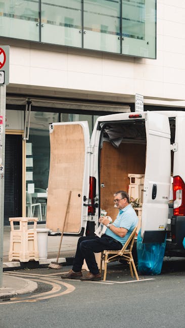 A man wearing glasses and a light blue shirt is seated on a wooden chair outside a white delivery van, which is parked on a city street fronting a modern building with large glass windows and white paneling. The van’s rear doors are open, revealing a transformation into a home relocation vehicle with visible packed cardboard boxes, wrapped furniture, and packing materials inside. A wooden panel leans against the van's side, and various packing supplies such as plastic wrap and blankets are seen in the process of being prepared for furniture transport. The man is holding and inspecting a small item, likely part of the packing process, as he rests beside the van. The street includes a traffic sign and a curb with a yellow line, indicating parking regulations, and the scene is set during daylight, illustrating a typical loading process for house removals carried out by Man and Van Harrow, supporting efficient packing and moving services for local relocations.