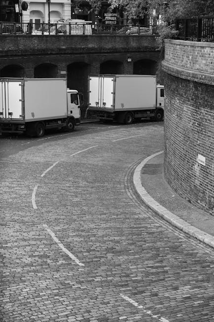 A black-and-white photograph of a cobbled street with three parked box trucks lined up along the curb, ready for a home relocation. The trucks are positioned parallel to a curved brick wall on the right side, which partially encloses the area outside a building with upper-level balconies visible in the background. The pavement shows dashed white lines and a circular marking for vehicle guidance. Inside the trucks, visible through open or partially open doors, are large cardboard boxes, wrapped furniture, and plastic packaging materials, indicating preparations for packing and moving household items. A trolley and securing straps can be seen near the trucks, suggesting the loading process. The scene appears to be in an urban residential area, with a focus on the logistics involved in furniture transport and house removals. Man and Van Harrow, a professional removals company, manages this moving operation, ensuring efficient and careful handling of items during the home relocation process.