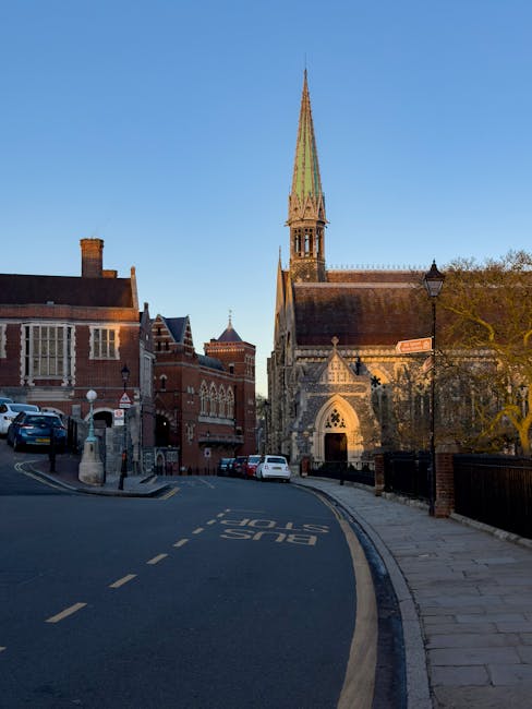 A street scene featuring a mix of historic and modern buildings in Harrow, with a prominent church tower with a tall, pointed spire made of stone and greenish roof tiles, situated behind a pavement and a narrow road marked with 'BUS STOP' in white paint. Several parked cars are lined along the street, and a lamppost is visible near the church entrance. The background shows a clear blue sky and trees with some foliage, indicating a bright day. This urban setting illustrates a typical area where house removals and furniture transport may occur, with key architectural features and street layout visible, reflecting the environment in which professional removals, such as those by Man and Van Harrow, operate.
