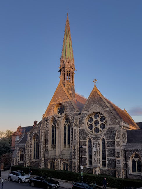 A view of a historic stone church with Gothic architectural features, including pointed arches, decorative circular windows, and a large central rose window, set against a clear blue sky during late afternoon light. The church has a tall, slender spire with a greenish patina, intricate masonry details, and a small cross atop the roof. In the foreground, a street scene shows parked cars, a narrow footpath, and some pedestrians. The church is situated on a street in Harrow, with neatly maintained hedges and visible urban surroundings. The image captures the exterior of the building, highlighting its architectural style and its prominent position within the local landscape, relevant for home relocation and furniture transport services provided by Man and Van Harrow, in the context of packing, moving, and loading processes typical of house removals in the area.