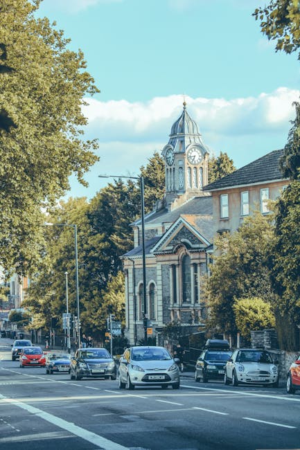 A street scene featuring a mix of historic and modern buildings in Harrow, with a prominent church tower with a tall, pointed spire made of stone and greenish roof tiles, situated behind a pavement and a narrow road marked with 'BUS STOP' in white paint. Several parked cars are lined along the street, and a lamppost is visible near the church entrance. The background shows a clear blue sky and trees with some foliage, indicating a bright day. This urban setting illustrates a typical area where house removals and furniture transport may occur, with key architectural features and street layout visible, reflecting the environment in which professional removals, such as those by Man and Van Harrow, operate.