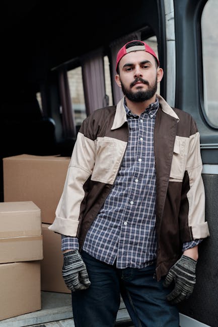A man with a beard, wearing a red cap backwards, a checkered shirt underneath a colour-blocked beige and brown jacket, stands inside the back of a moving van. The van's interior is visible, showing several cardboard boxes of varying sizes stacked on the floor, secured for transport. The man is wearing work gloves and appears to be involved in the home relocation process, possibly preparing to load or unload furniture and packing materials. The environment suggests an organised loading process typical of furniture transport or house removals, supported by the professional context of Man and Van Harrow, as indicated by the webpage title and related services for tight access jobs like those at Pinner Park Estate. The lighting is natural, and the focus is on the man and the items around him, emphasizing the logistics of packing and moving homes.