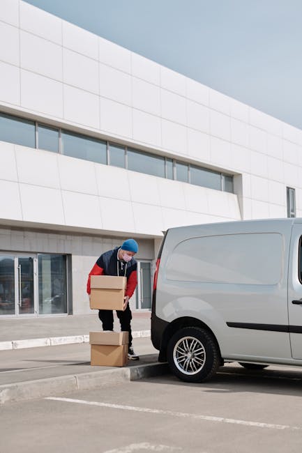 A man wearing a blue beanie, red and navy jacket, and black trousers is lifting a cardboard box from a stack of two boxes positioned on paved ground outside a modern white building with large glass doors. He appears to be preparing to load the box into a light grey van parked nearby, which features a sliding side door and black wheel arches. The vehicle is situated close to the building's entrance, likely part of a home relocation or furniture transport process. The scene is set in daylight, with clear weather and natural lighting illuminating the scene, emphasizing the activity of packing and moving goods as part of professional removals services, such as those offered by Man and Van Harrow, aligning with the context of tight access jobs at Pinner Park Estate.