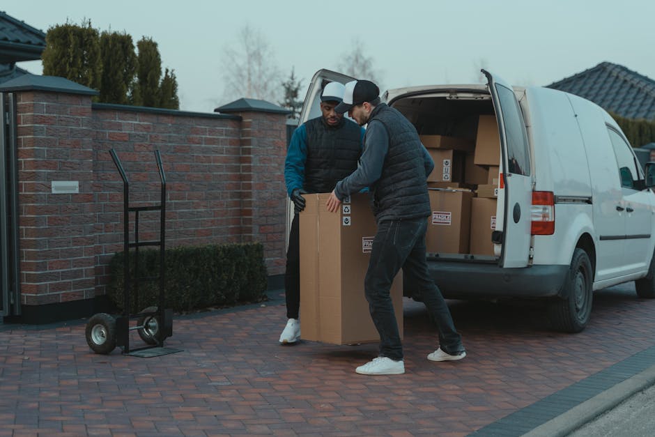 A man with a beard, wearing a red cap backwards, a checkered shirt underneath a colour-blocked beige and brown jacket, stands inside the back of a moving van. The van's interior is visible, showing several cardboard boxes of varying sizes stacked on the floor, secured for transport. The man is wearing work gloves and appears to be involved in the home relocation process, possibly preparing to load or unload furniture and packing materials. The environment suggests an organised loading process typical of furniture transport or house removals, supported by the professional context of Man and Van Harrow, as indicated by the webpage title and related services for tight access jobs like those at Pinner Park Estate. The lighting is natural, and the focus is on the man and the items around him, emphasizing the logistics of packing and moving homes.